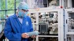 Een persoon in een blauwe Unilever-labjas, haarnetje, veiligheidsbril en mondkapje onderzoekt een grijze plastic fles in een fabrieksomgeving, met industriële apparatuur op de achtergrond.